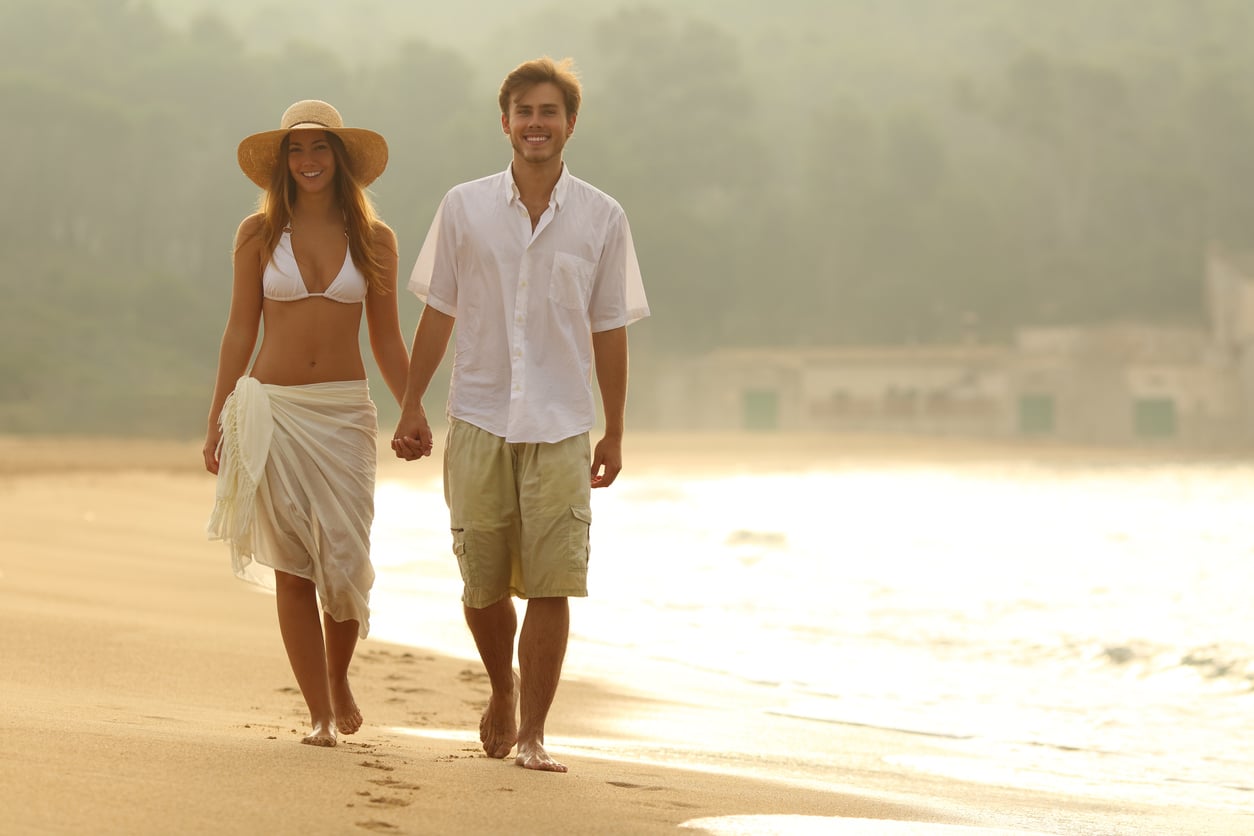 A smiling couple walks hand in hand along a sandy beach at sunset, enjoying their beach vacation savings. The woman wears a white bikini, sarong, and sunhat, while the man wears a white shirt and shorts. The ocean and trees are visible in the background.