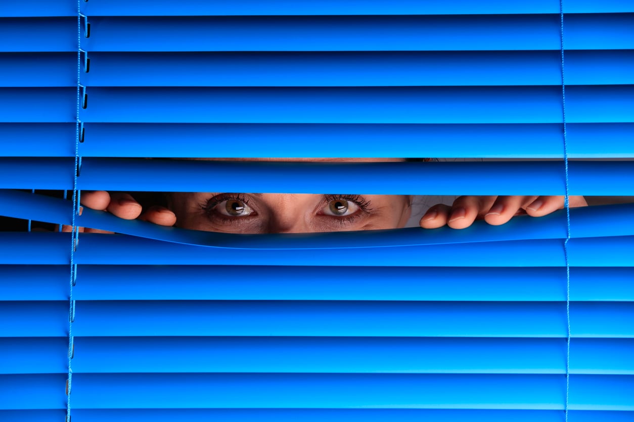 A person peeks through the gap of closed blue window blinds, with only their eyes and hands visible—a quiet moment that evokes the curiosity and stillness of life before smartphones.