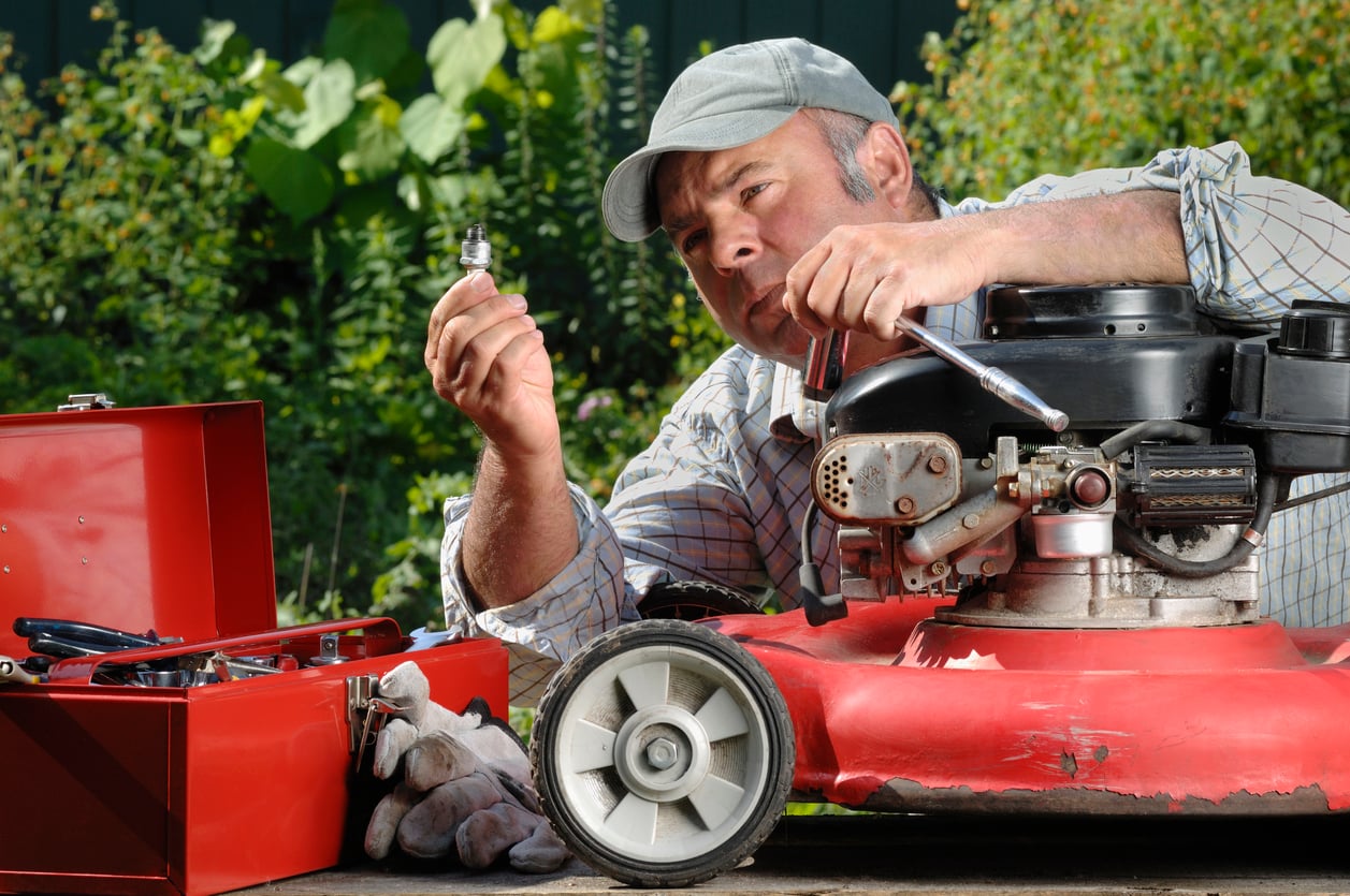 A man in a cap repairs a red lawnmower outdoors, examining a spark plug while holding a wrench. Various products, including a red toolbox and work gloves, are next to him on the table. Greenery is visible in the background.