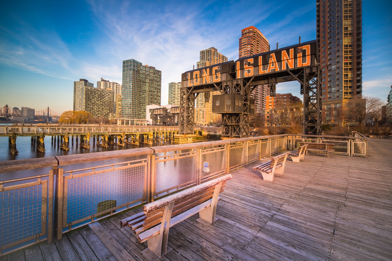 A waterfront boardwalk with benches overlooks the iconic Long Island sign and high-rise buildings in Long Island City, Queens, New York, under a clear blue sky at sunset.