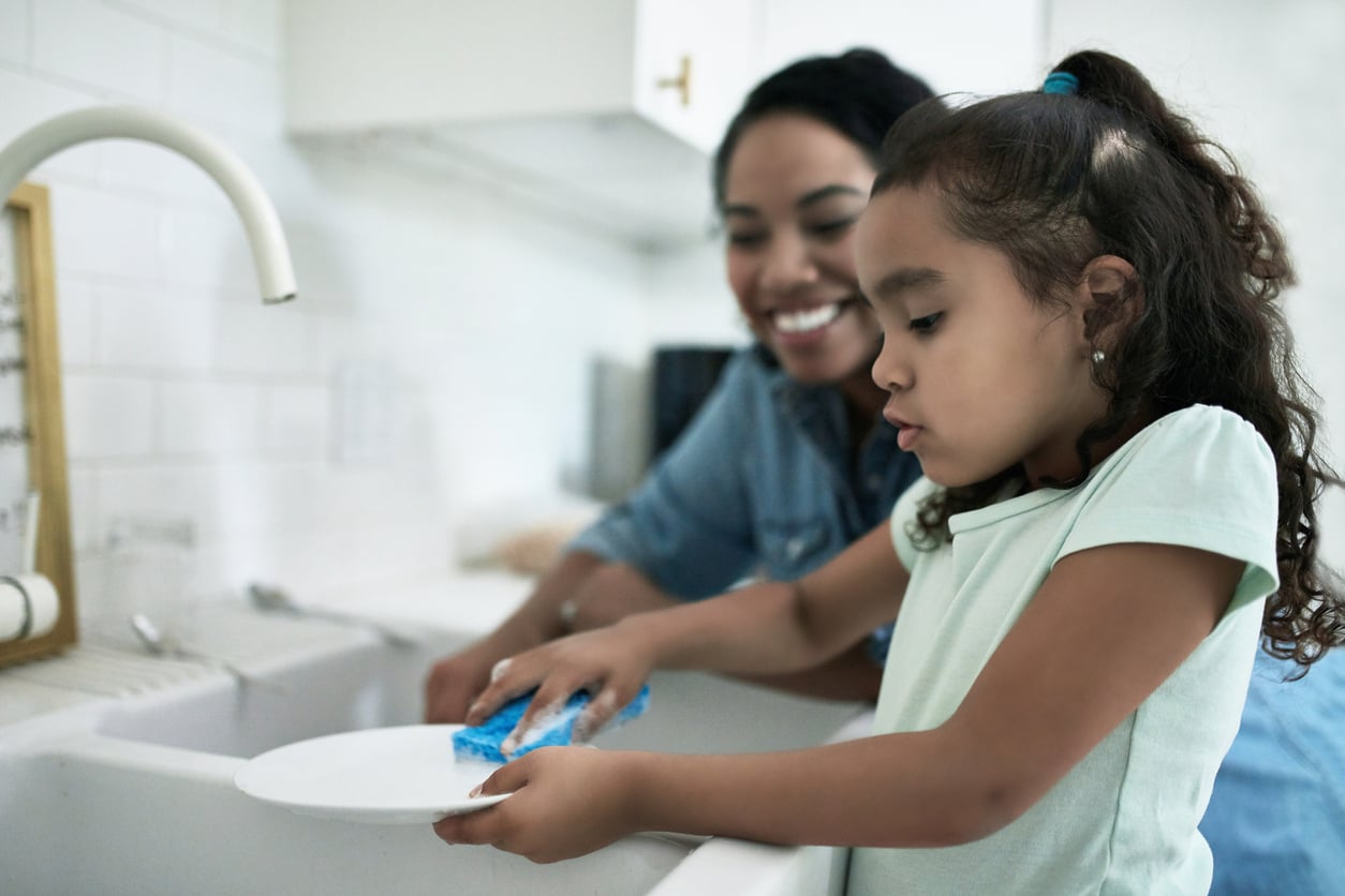 A young girl washes a plate at a kitchen sink with a sponge while an adult woman smiles and watches beside her.