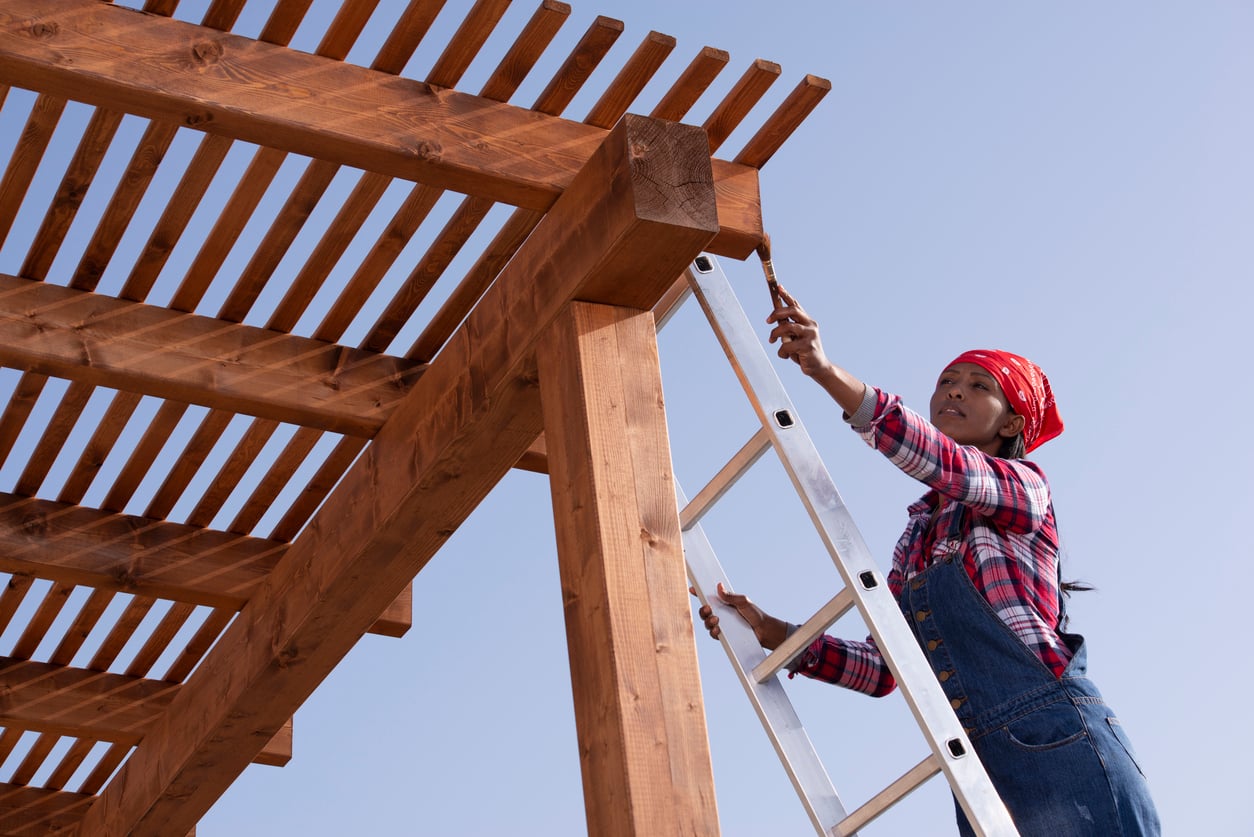 A person wearing overalls and a red bandana stands on a ladder, working on a wooden pergola—one of the many quality outdoor home products—under a clear blue sky.