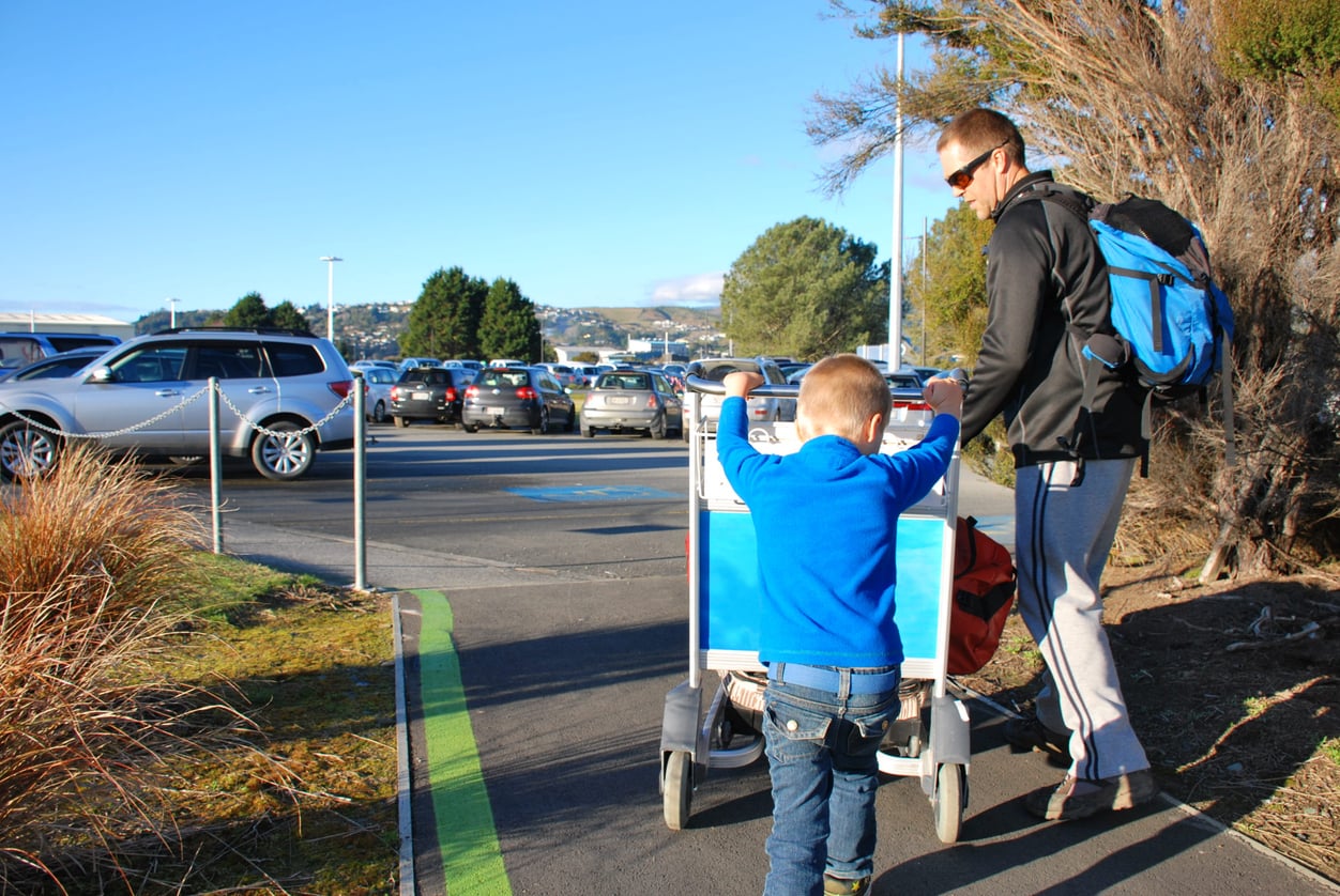 A man and a young boy push an airport luggage cart along a sidewalk near a parking lot on a sunny day, with cars and trees in the background, perhaps beginning their journey after arranging vacation swaps.