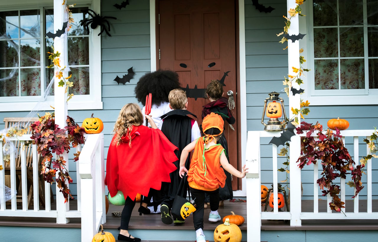 Four children in Halloween costumes approach a decorated porch with pumpkins, leaves, bats, and a jack-o’-lantern. The kids are trick-or-treating—a classic Halloween activity—at a house adorned with festive autumn and Halloween decorations.