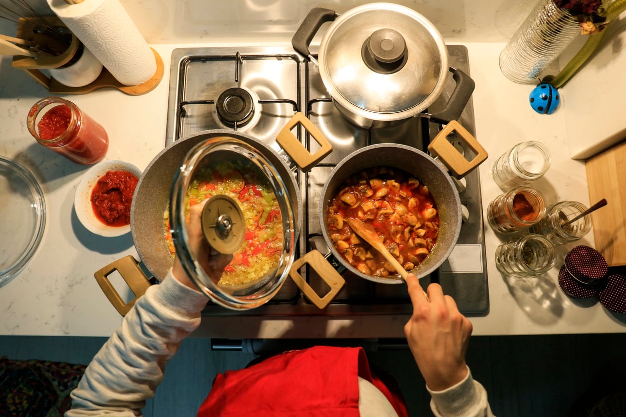 Person cooking on a stove, stirring a pot of vegetables with a wooden spoon while holding a lid over another pot. Jars, sauce, and kitchen utensils are nearby on a white countertop.
