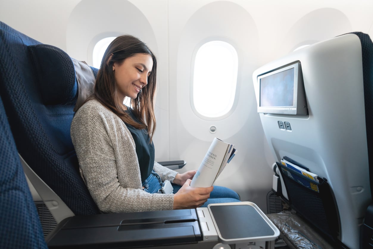 Airline perks. A woman sits in an airplane seat by the window, smiling while reading a magazine or book. She is wearing a gray cardigan and jeans, and there is an in-flight entertainment screen in front of her.