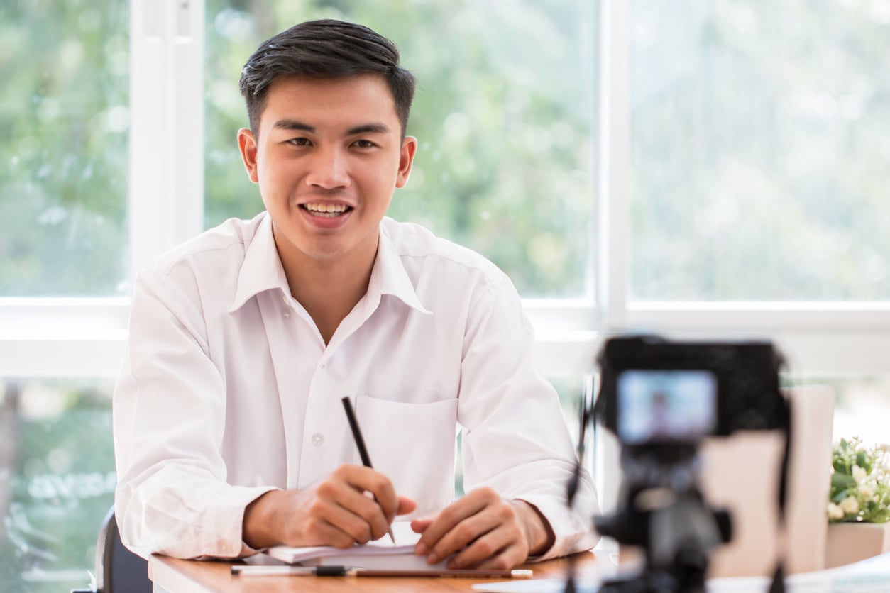 A young man in a white shirt sits at a desk, smiling and holding a pen, with a notebook in front of him. A camera on a tripod is positioned in the foreground, recording him. Bright windows and greenery are in the background.