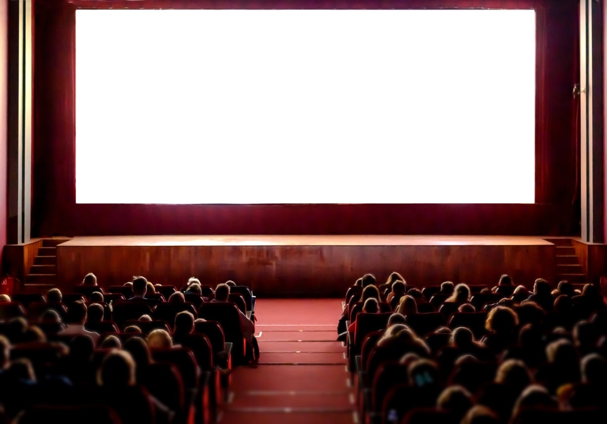 A large group of people sit in a dark movie theater facing a bright, blank white screen. The red seats fill up as anticipation builds—everyone’s eager for the film and maybe to try out some movie theater hacks during the show.