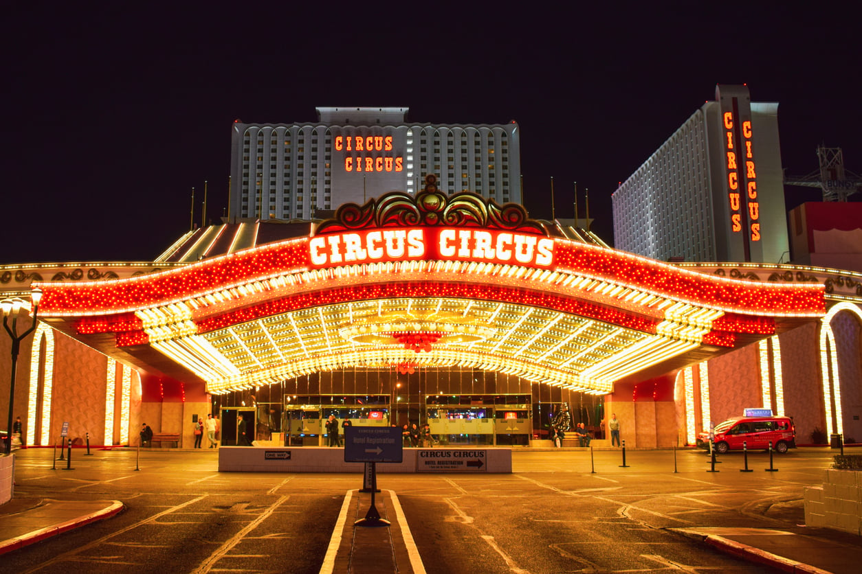 Circus Circus casino entrance at night, lit by bright yellow and red lights, with a large illuminated sign displaying “Circus Circus” and several cars parked in front.