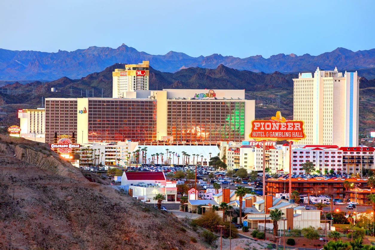 View of several brightly lit casino hotels in Laughlin, Nevada at dusk, with colorful neon signs, palm trees, parking lots, and mountains in the background under a clear sky—showcasing one of the most affordable travel destinations for an exciting getaway.