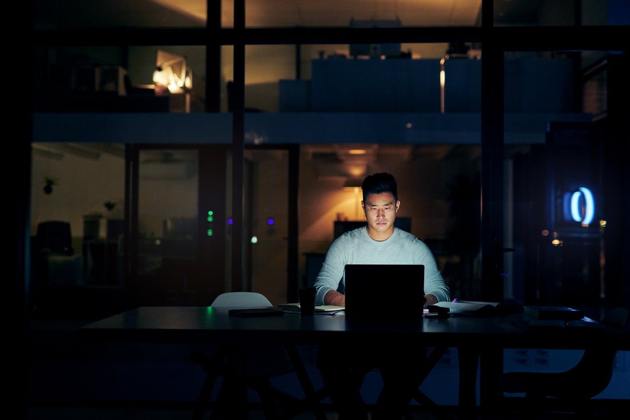 A man sits alone at a desk in a dark office, illuminated by his laptop screen as he works late into the night—a quiet reflection of how families in America are having fewer children. Papers and a coffee cup rest on the table, with office lights glowing softly behind him.