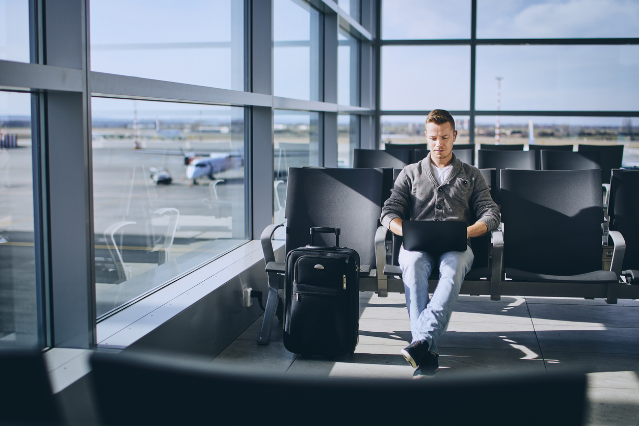 A man sits in an airport terminal waiting area, working on a laptop with a suitcase beside him. Large windows show airplanes and the runway outside on a sunny day.