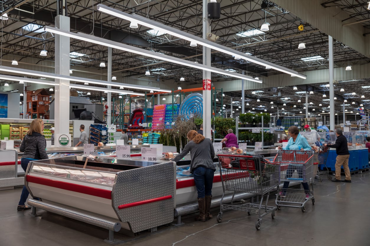 Shoppers with carts browse items in the refrigerated section of a large, well-lit warehouse store with high ceilings and various products on display.