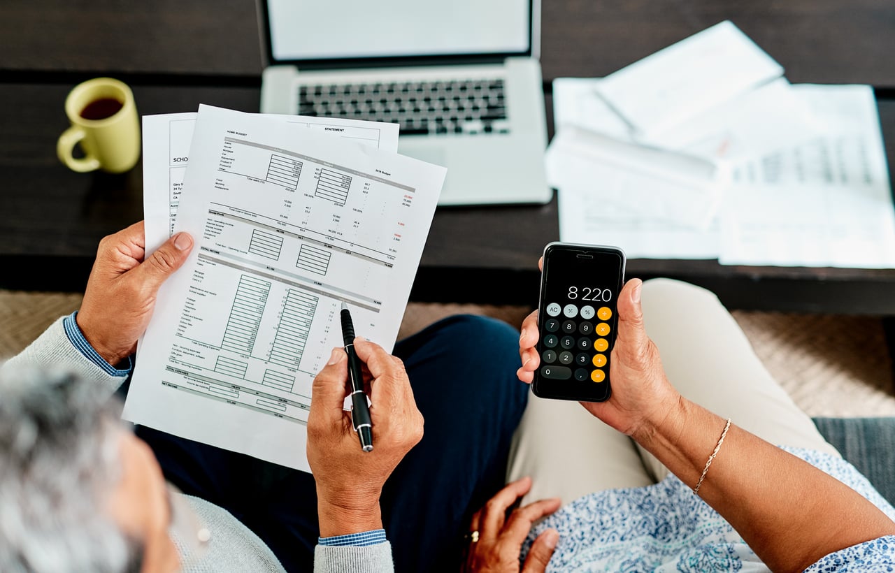 Two people sit at a table reviewing financial documents and using a smartphone calculator. A laptop and papers are on the table, and one person holds a pen while the other holds the phone showing "8,220.