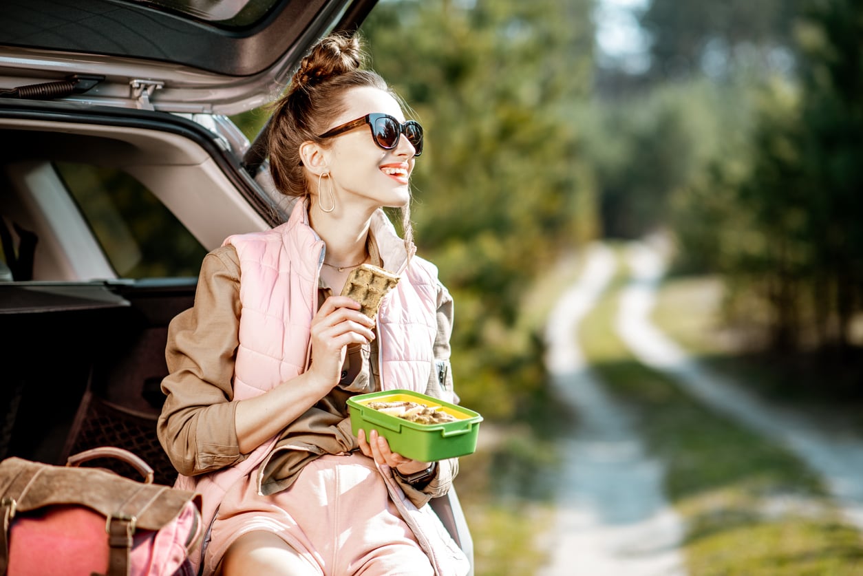 A woman in sunglasses sits in the open trunk of a car, eating a sandwich and holding a green lunchbox. She smiles, surrounded by trees on a sunny day. A dirt path extends into the forest.