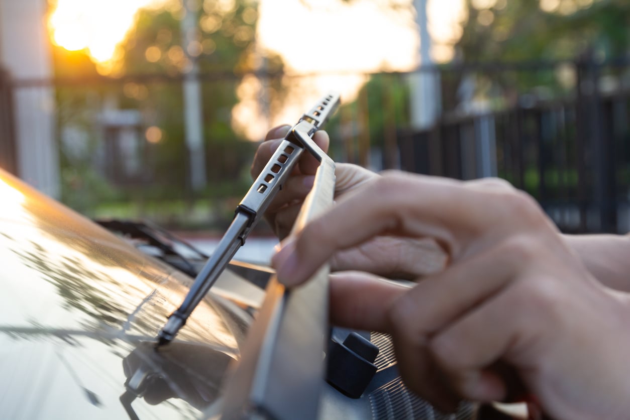Close-up of a person’s hands installing or adjusting a windshield wiper blade on a car, with sunlight and a blurred outdoor background—showcasing smart car maintenance hacks in action.