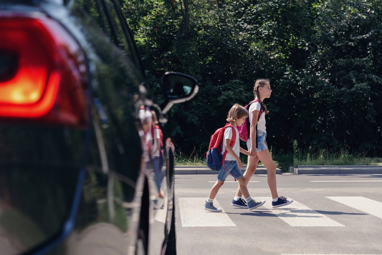 Two children with backpacks walk across a crosswalk while a car waits nearby. Trees and greenery are visible in the background on a sunny day.