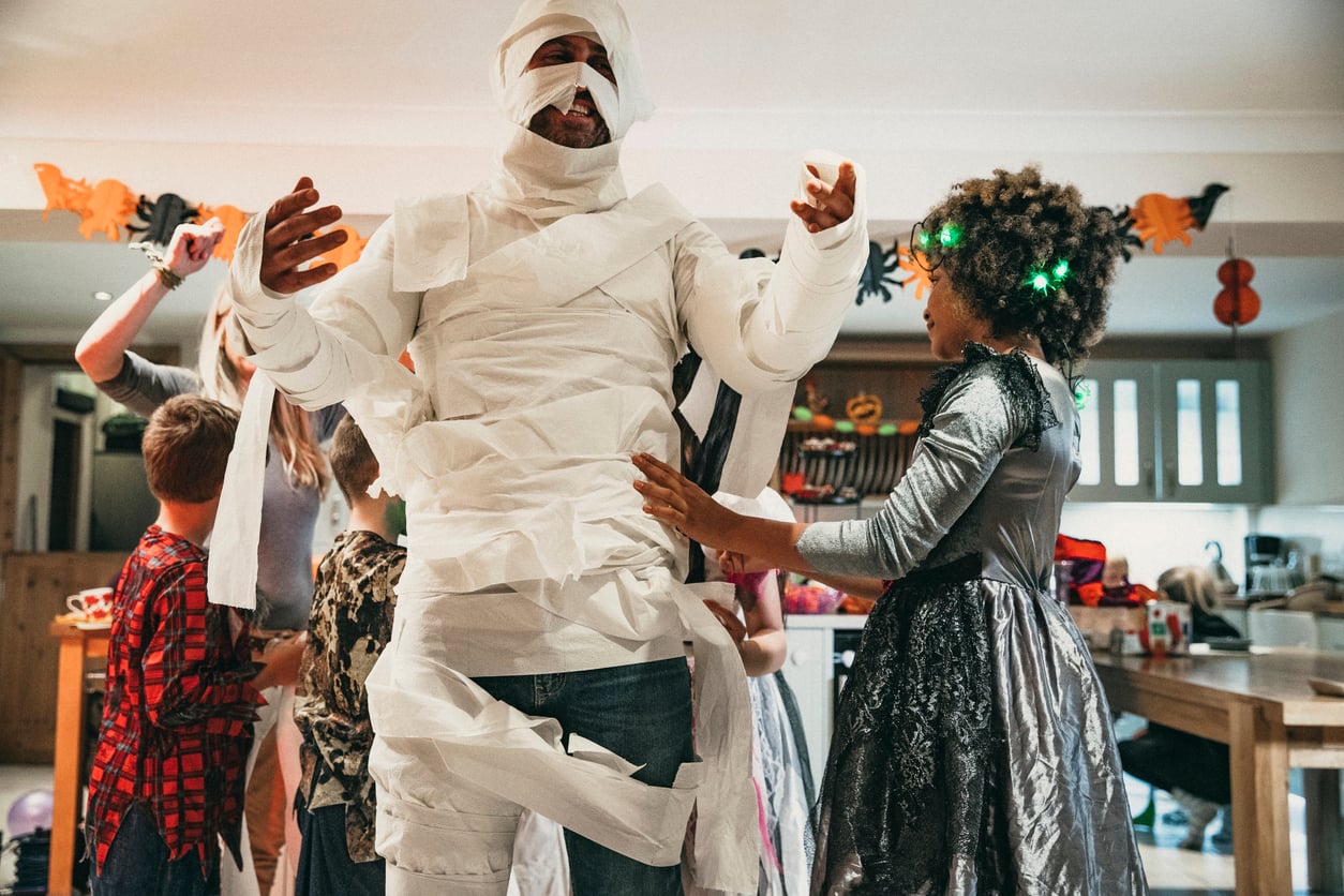 A person wrapped in toilet paper like a mummy stands in a kitchen, surrounded by children in Halloween costumes, laughing and having fun during a party filled with exciting Halloween activities. Decorations are visible in the background.
