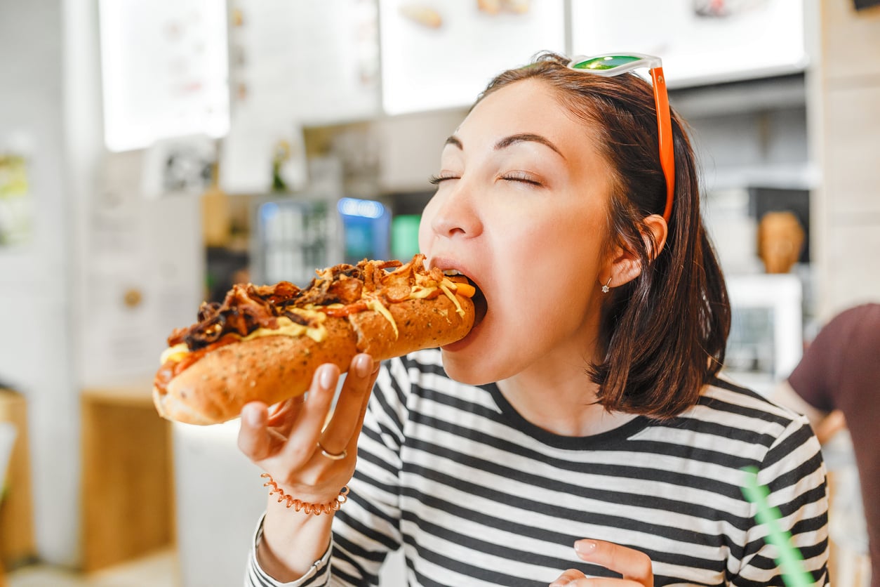 A woman in a striped shirt with sunglasses on her head is about to take a big bite of a loaded hot dog, sitting in a casual restaurant.