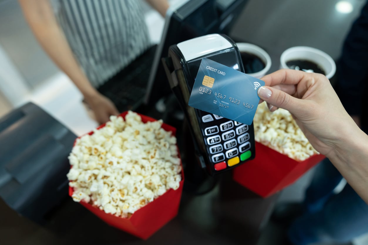 A person holds a credit card over a payment terminal to make a contactless payment at a counter with two red tubs of popcorn and two drinks—one of those movie theater hacks for quick and easy snacks before the film starts.