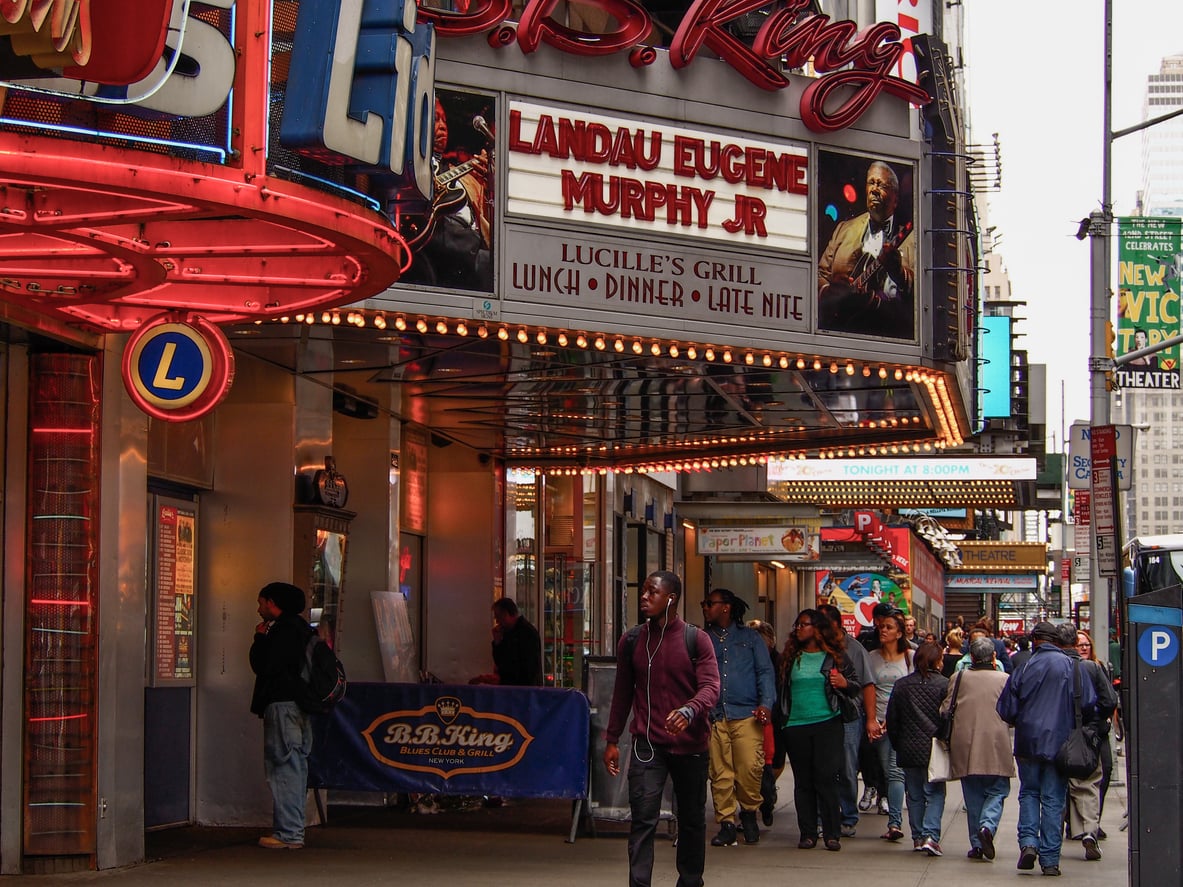 People walk past the B.B. King Blues Club in New York City, where a marquee advertises a performance by Landau Eugene Murphy Jr. Neon signs and colorful lights decorate the lively street scene, attracting visitors who are mindful of vacation costs as they soak in the vibrant atmosphere.