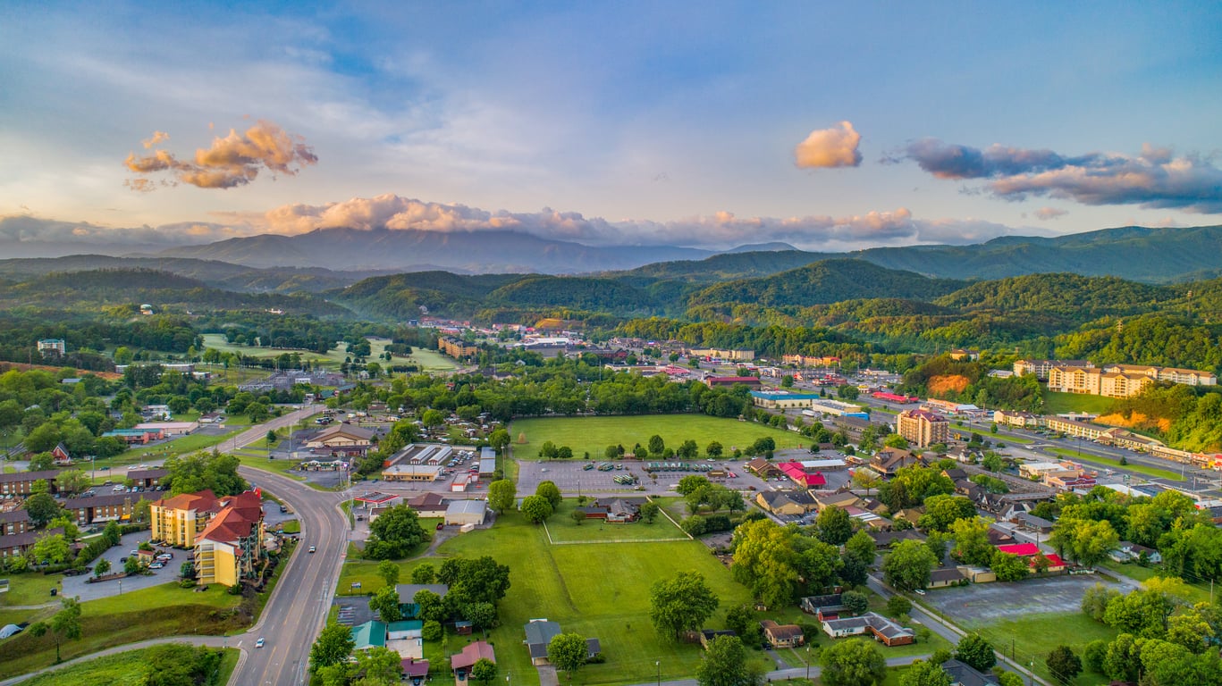 Aerial view of a small town with buildings, roads, and fields, surrounded by green hills and distant mountains under a blue sky with scattered clouds at sunset.