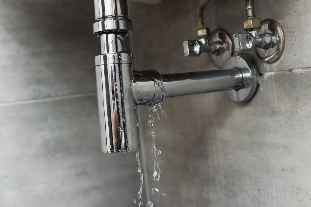 A close-up of a leaking chrome pipe under a sink, with water dripping onto a gray tiled wall—highlighting a plumbing issue that could increase household expenses.