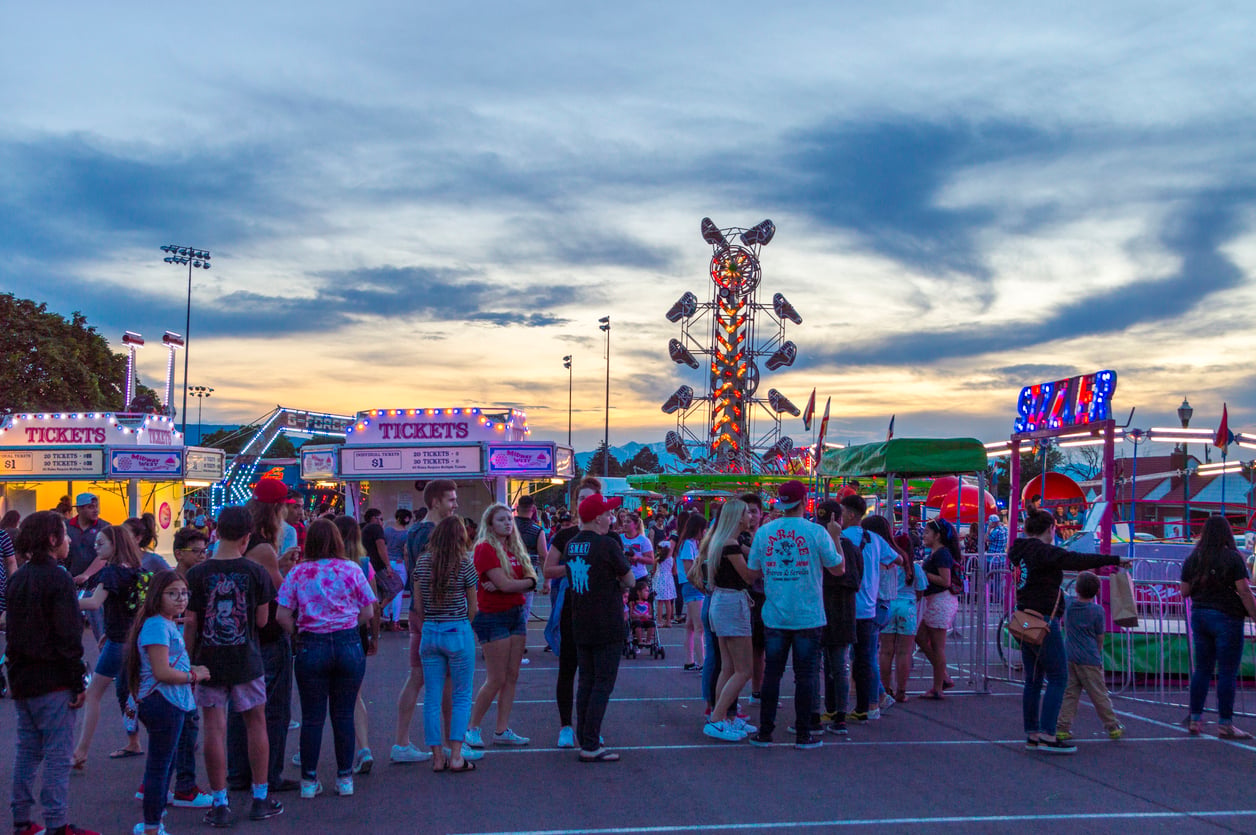 People wait in line at a lively outdoor carnival at sunset, with colorful ticket booths, rides, and lights in the background. The sky is partly cloudy, adding to the festive atmosphere.
