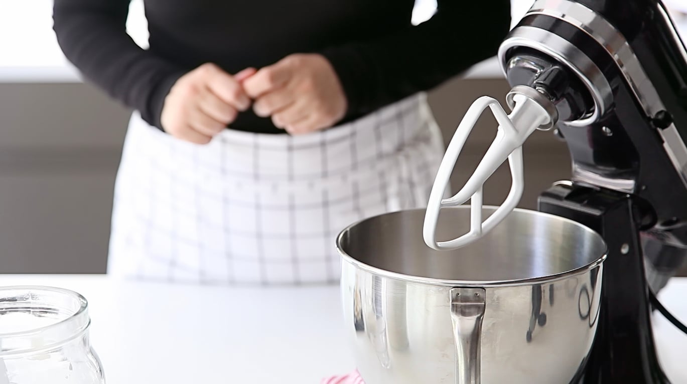 A person wearing a black shirt and a white checkered apron stands behind a stand mixer with a paddle attachment in a modern kitchen, ensuring quality over products that don't last. Their hands are clasped, and a glass jar is visible on the counter.