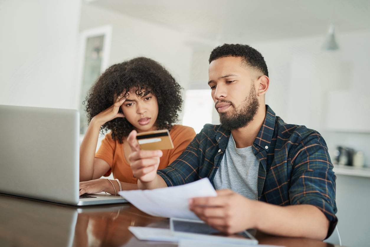 A concerned couple sits at a table with a laptop, looking at a credit card and paperwork, appearing worried about their finances.
