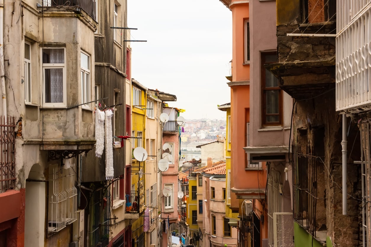 Narrow street lined with colorful, weathered buildings featuring balconies and satellite dishes, laundry hanging outside, and a distant view of a cityscape under a cloudy sky—reminding travelers that discovering authentic local neighborhoods is one of the joys you miss when making common hotel booking mistakes.