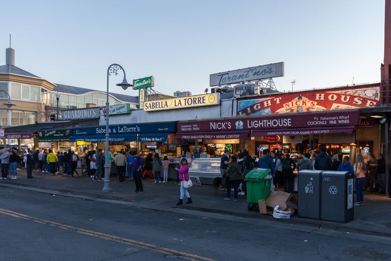 A busy street scene at Fisherman's Wharf in San Francisco, with crowds gathered outside popular seafood restaurants and colorful signs, including Sabella & La Torre and Nick's Lighthouse, at dusk.