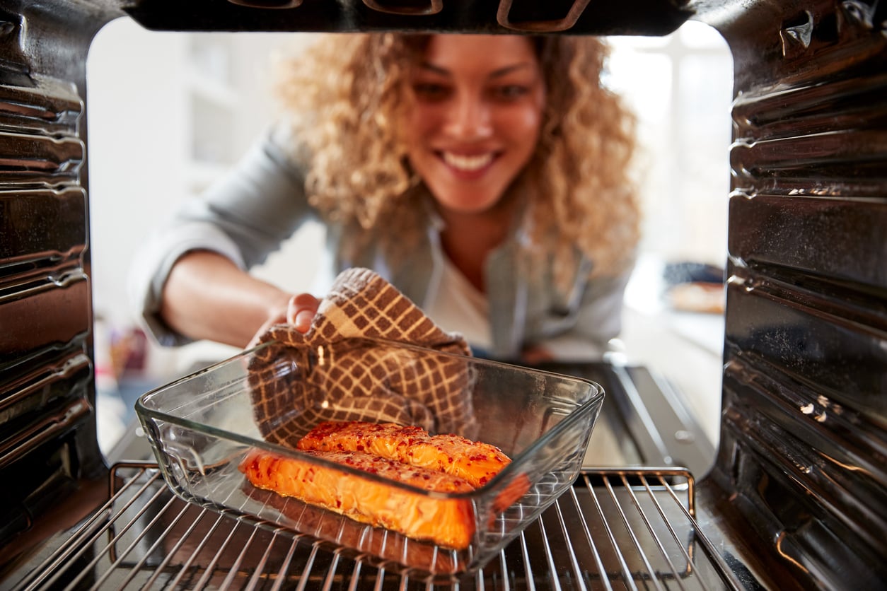 A woman smiling as she uses an oven mitt to remove a glass baking dish with cooked salmon from an oven, viewed from inside the oven.