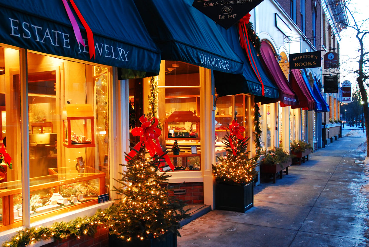 A row of warmly lit shop windows decorated with holiday lights, red bows, and small Christmas trees lines a snowy evening sidewalk—capturing the magical charm found in the best Christmas towns in America.