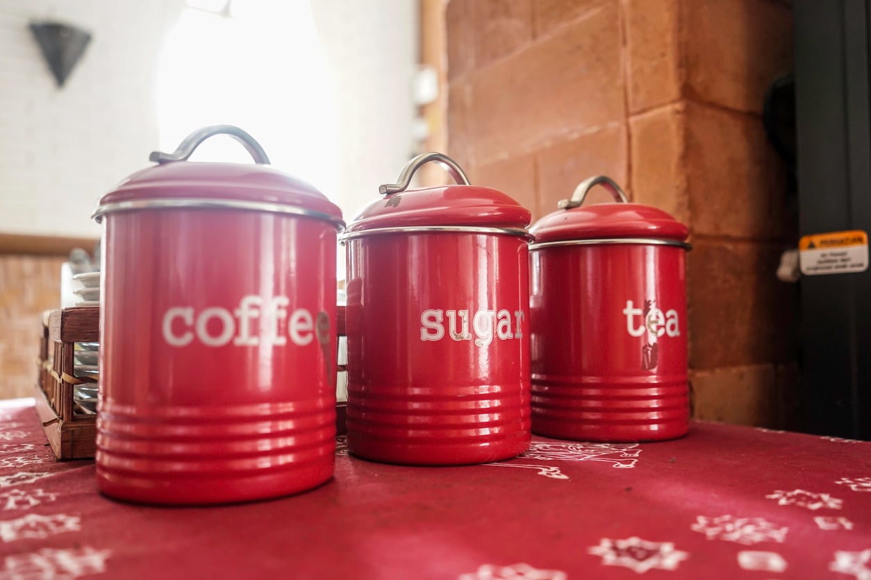 Three red canisters labeled "coffee," "sugar," and "tea" sit on a red patterned tablecloth among other charming kitchen items in a cozy kitchen setting, with sunlight coming through a window in the background.