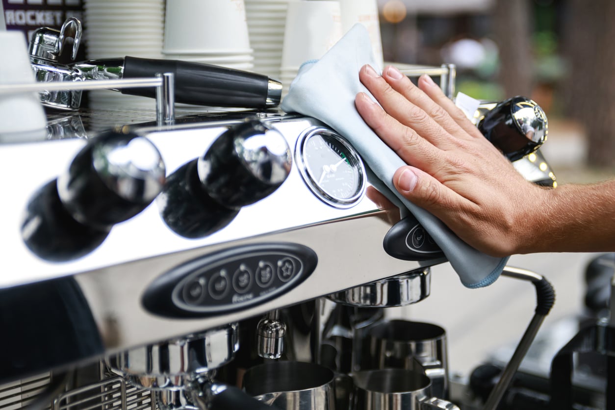 A person wipes the shiny surface of an espresso machine with a cloth, surrounded by stacked cups, coffee-making tools, and other products essential for crafting the perfect brew.