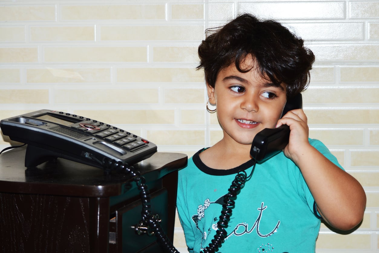 A young child with short dark hair smiles while holding a black corded phone to their ear, standing next to a desk with a phone, against a tiled wall background.