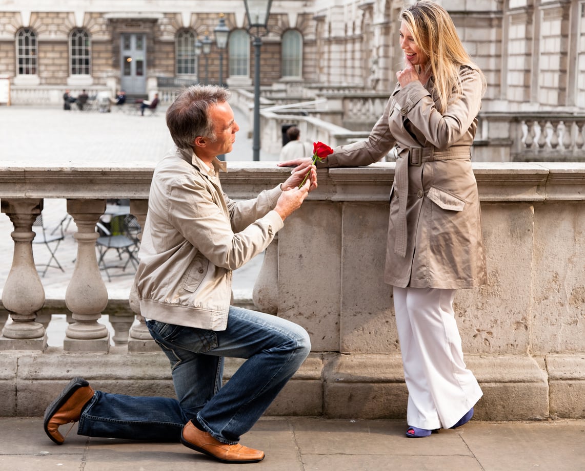 A man kneels on one knee, offering a red rose to a smiling woman leaning against a stone railing in an urban setting—a romantic proposal at a time when families in America are having fewer children.