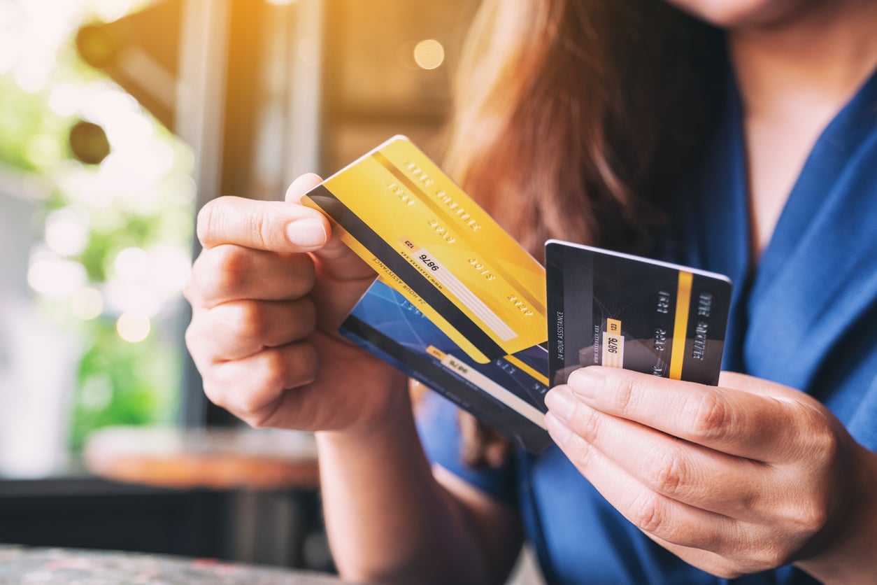 A person wearing a blue shirt holds two credit cards, one gold and one black, inside a bright room with sunlight shining through the window in the background, perhaps preparing to manage their monthly bills.