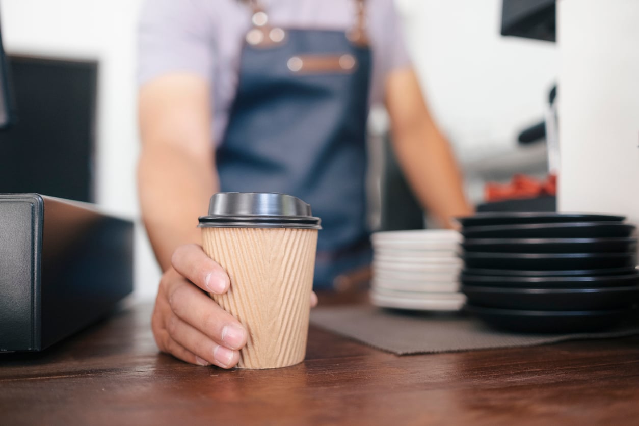 A barista wearing a blue apron hands a takeaway coffee cup with a black lid across a wooden counter, near stacked plates and a payment terminal—reminding customers how small treats can add up in their household expenses.