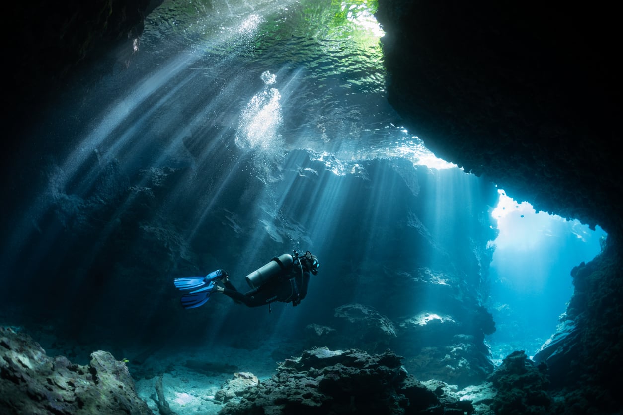 A scuba diver indulges in one of the world’s most expensive hobbies as they swim through an underwater cave, with sunlight streaming down through openings above and illuminating rocks and clear blue water.