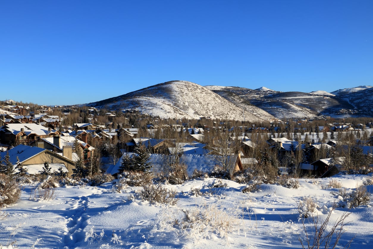 Snow-covered houses and trees, reminiscent of quaint American towns, sit at the base of a rounded hill under a clear blue sky, with footprints visible in the foreground snow.