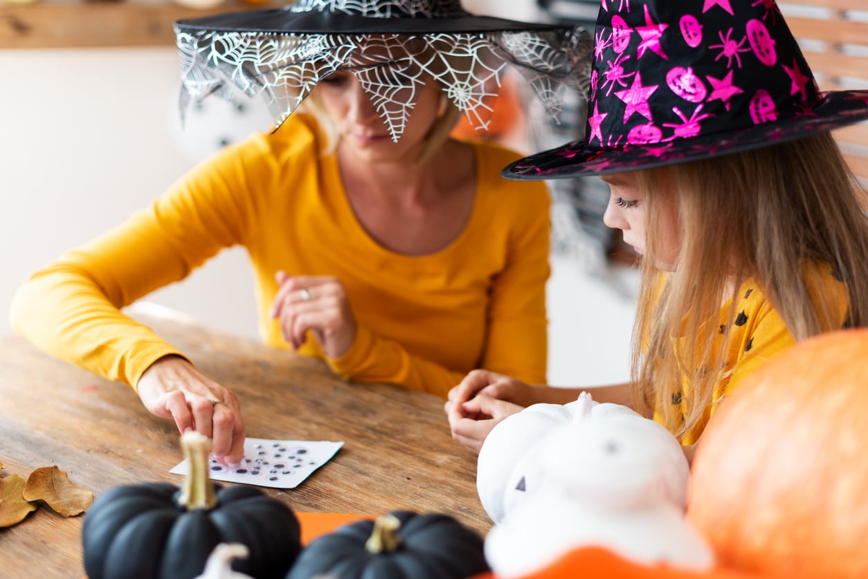 Two people in Halloween hats and orange shirts enjoy Halloween activities as they play cards at a wooden table decorated with pumpkins and painted gourds.
