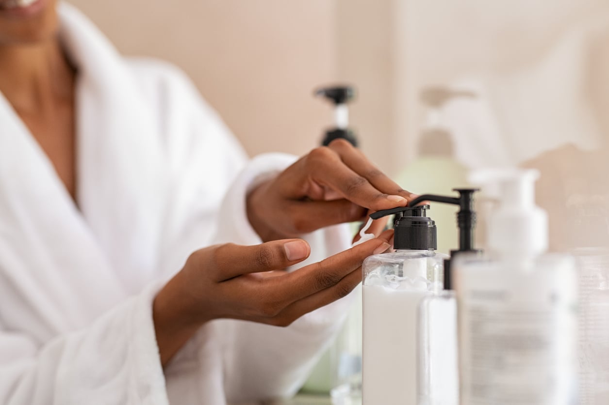 A person in a white bathrobe pumps lotion or soap from a dispenser onto their hand, with additional bottles visible on the counter.
