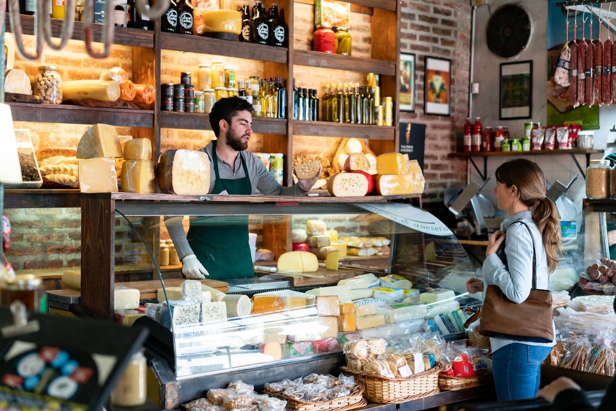 A man behind a cheese counter serves a woman in a shop filled with various cheeses, jars, and loaves of bread—an ideal stop for travelers enjoying vacation swaps. The shop has brick walls and wooden shelves displaying products, creating a warm, inviting atmosphere perfect for discovering local specialties.