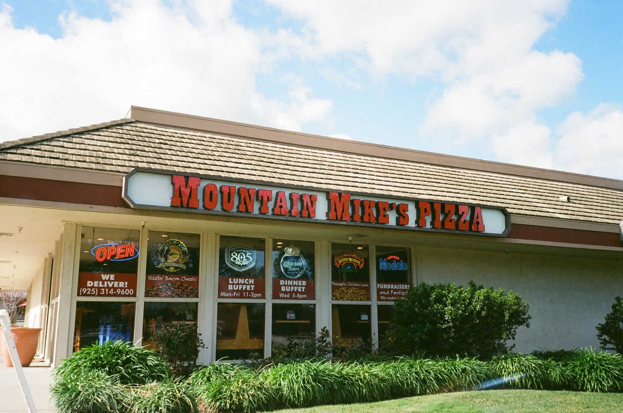 Exterior of Mountain Mike’s Pizza restaurant, a standout among regional fast-food chains, with a red sign, windows displaying posters, and an “OPEN” sign. Green shrubs line the front under a partly cloudy sky.