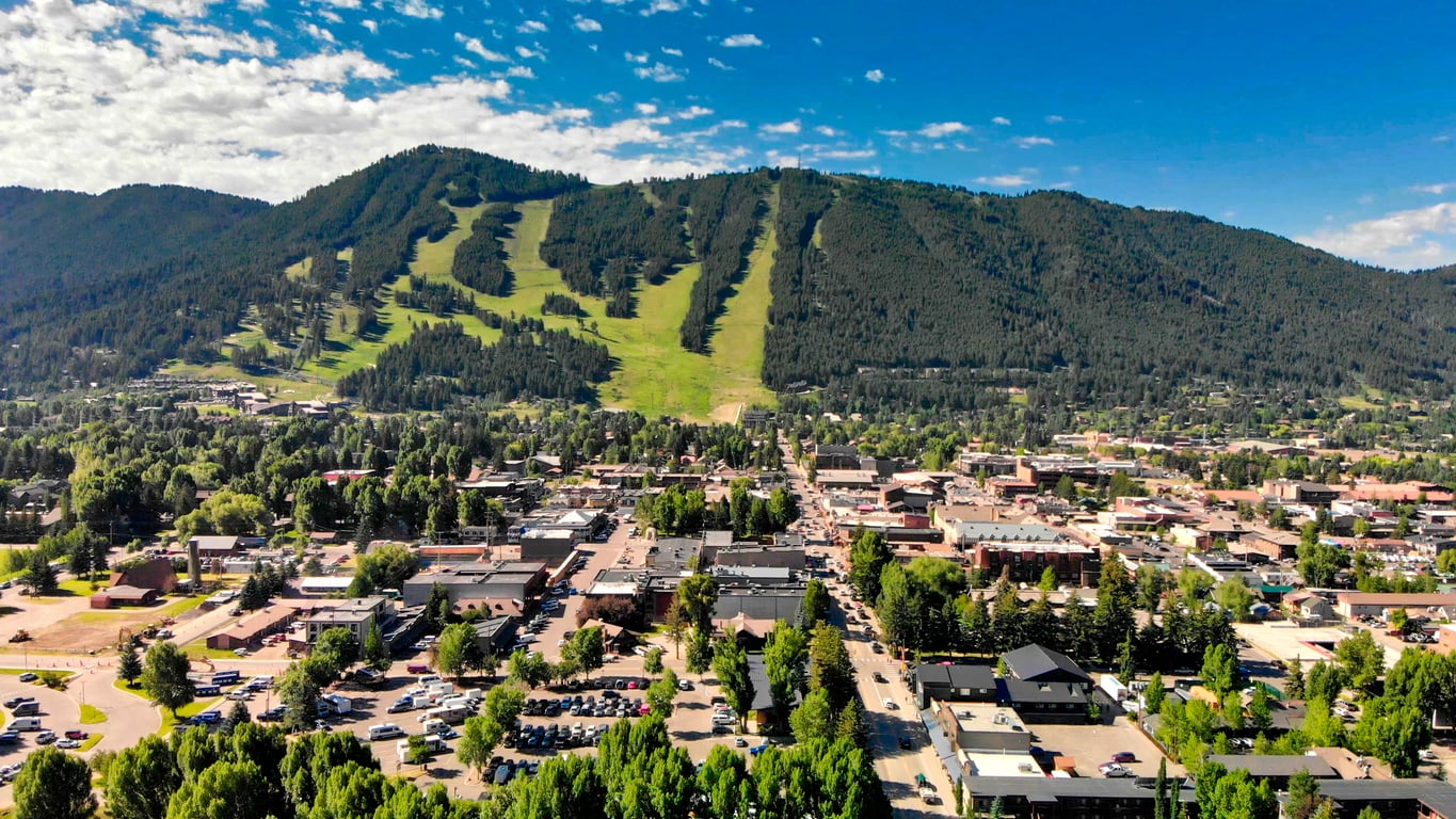 Aerial view of a small town with buildings and streets nestled at the base of a forested mountain with grassy ski slopes under a blue sky with scattered clouds, making it one of the charming travel destinations for nature lovers and adventure seekers alike.