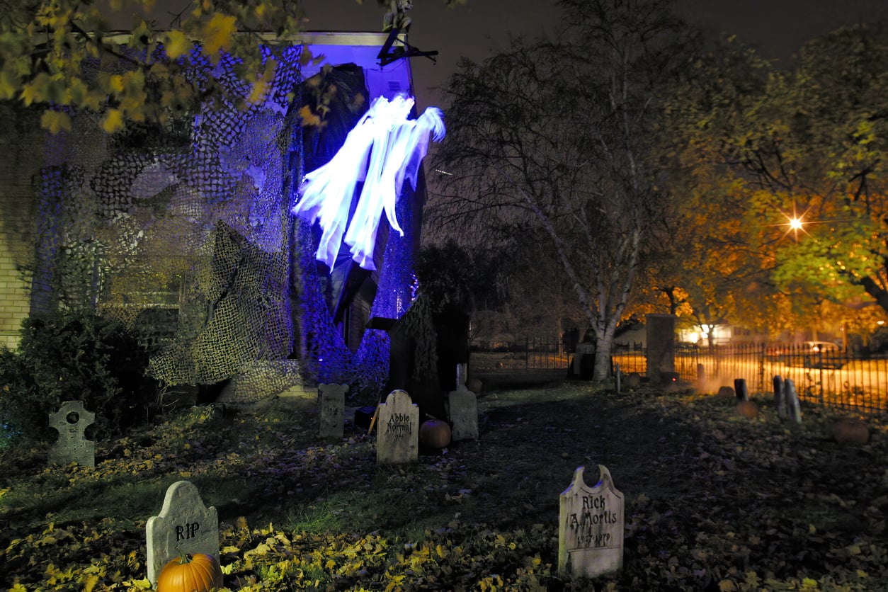 A spooky Halloween yard at night features glowing ghost decorations, several tombstones, fallen leaves, and trees with orange streetlights in the background, creating a classic Halloween atmosphere.