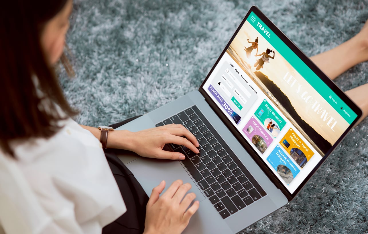 A woman sits on a gray carpet using a laptop displaying a travel booking website with destination packages and a "Let’s Go Travel" banner, carefully reviewing her options before booking a flight.