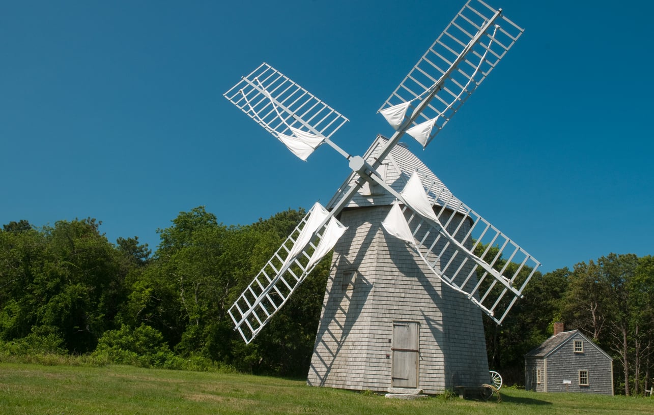 A traditional wooden windmill with white sails stands on green grass, reminiscent of those found in charming beach towns, with a small house and trees in the background under a clear blue sky.